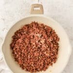 A white skillet containing cooked ground beef and diced onions on a light-colored surface.