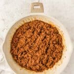 Cooked ground beef with sauce in a white skillet, viewed from above on a light-colored surface.