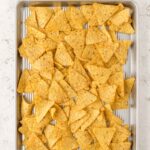 A metal baking tray filled with scattered yellow corn tortilla chips on a light stone surface.