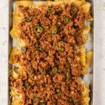 Sheet pan with nachos topped with ground meat, shredded cheese, and sliced jalapeños on a light-colored surface.
