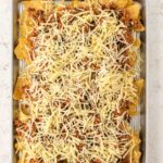 A baking sheet topped with tortilla chips, cooked ground meat, and shredded cheese, ready to be baked.