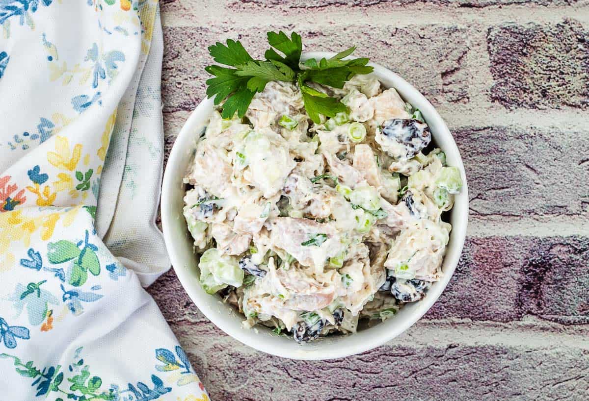 A bowl of chicken salad with celery, grapes, and parsley garnish sits on a brick-patterned surface next to a floral napkin, offering fresh twists on classic Chicken Salad Recipes.