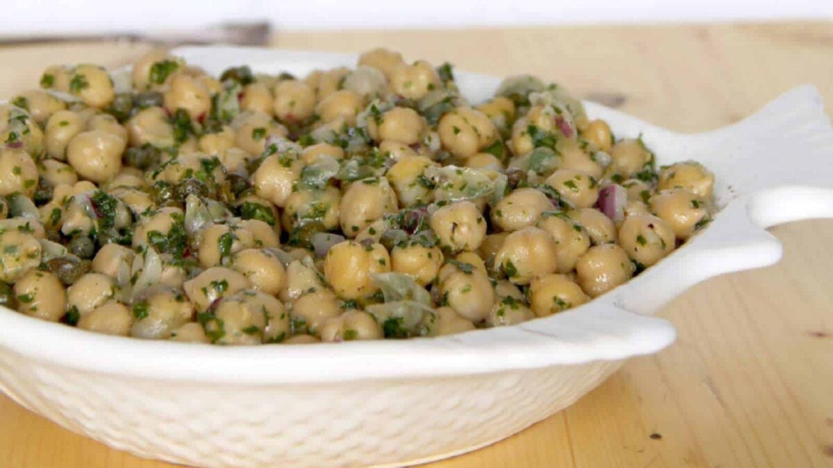 A white fish-shaped dish filled with chickpea salad mixed with chopped herbs and vegetables sits on a light wooden surface—one of the most satisfying chickpea dishes for those who love filling meals.