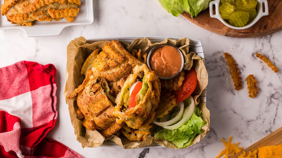 A basket with a fried chicken sandwich, lettuce, tomato, onion, waffle fries, and dipping sauce sits on a marble surface—perfect for those seeking delicious dinner recipes or craving creative cheeseburger ideas. Pickles and a red checkered napkin are nearby.