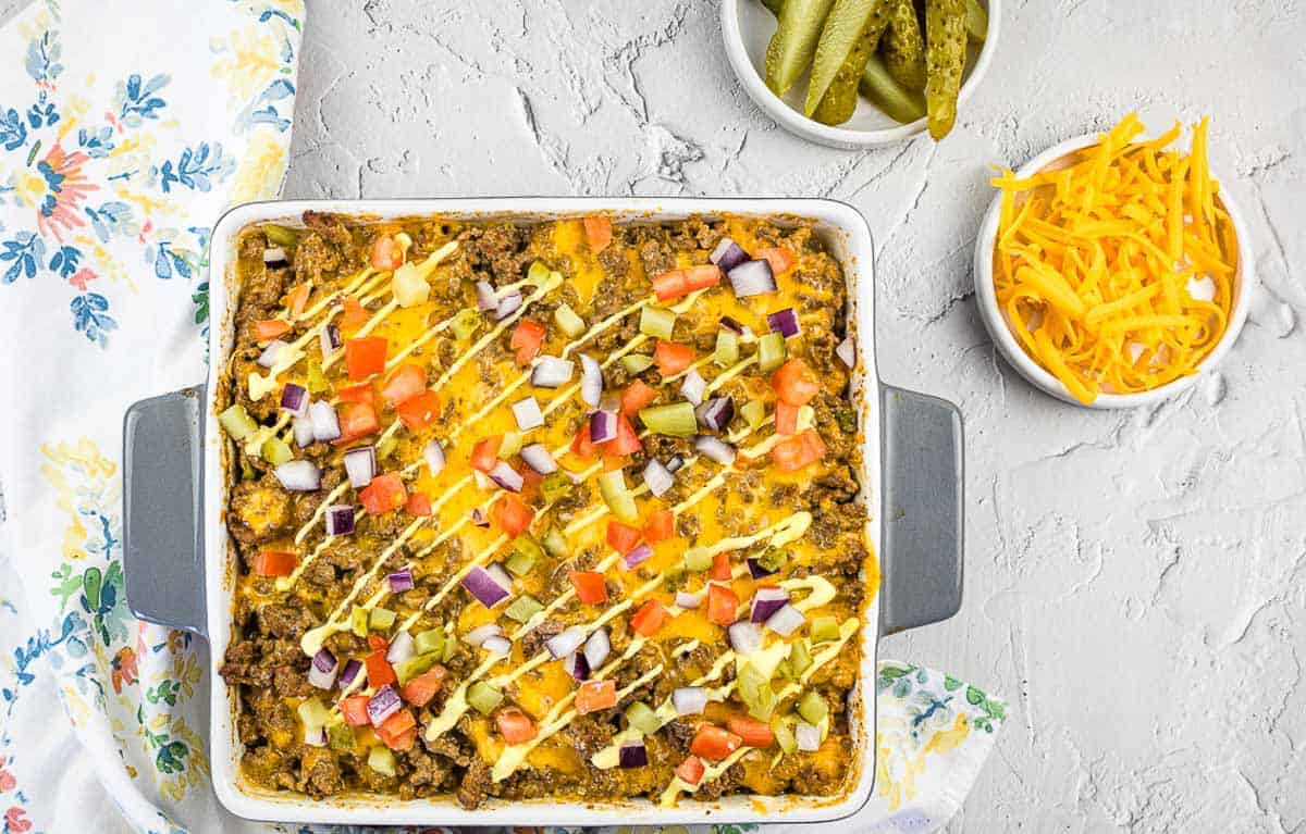 A square casserole dish filled with baked cheeseburger casserole, topped with chopped tomatoes, onions, pickles, and drizzled sauce—perfect for those seeking creative cheeseburger ideas or easy dinner recipes. Shredded cheese and pickles are in bowls nearby.