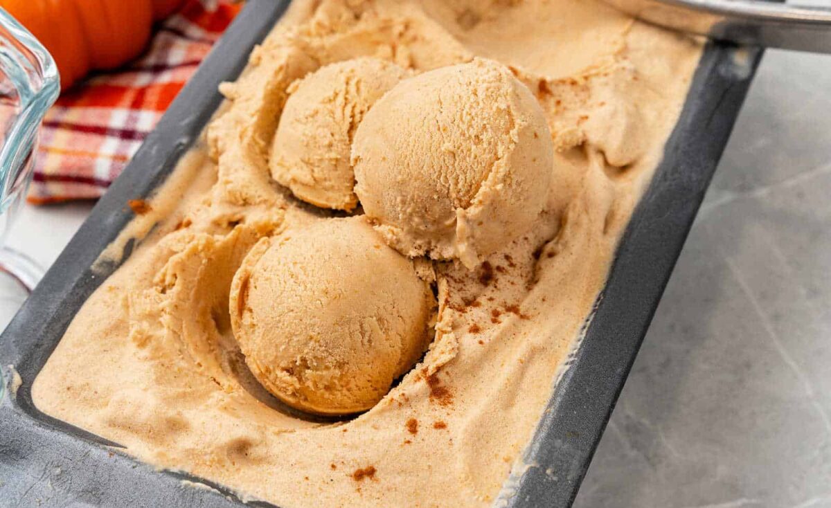 Three scoops of creamy, light brown ice cream desserts rest in a metal loaf pan, topped with a sprinkle of spice and set against a plaid cloth backdrop.