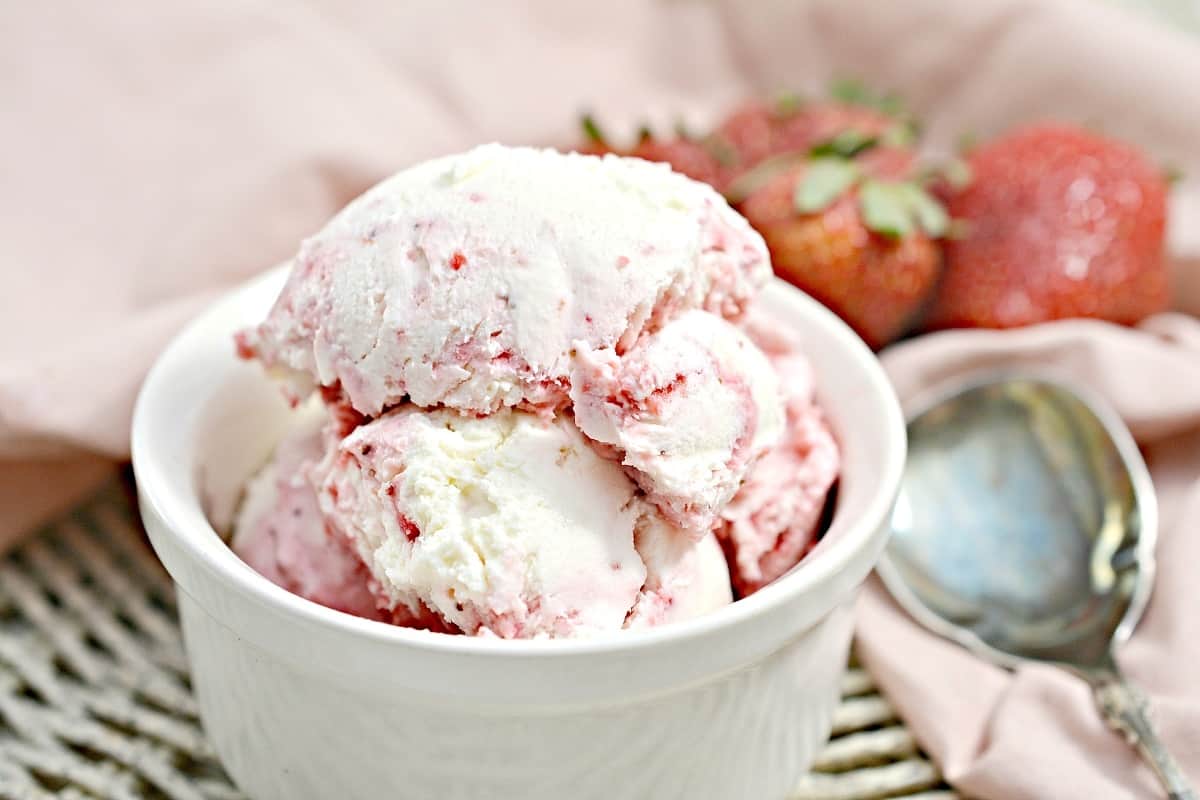 A white bowl filled with scoops of strawberry ice cream sits next to a spoon and fresh strawberries on a light pink cloth—perfect for inspiring new dessert ideas.