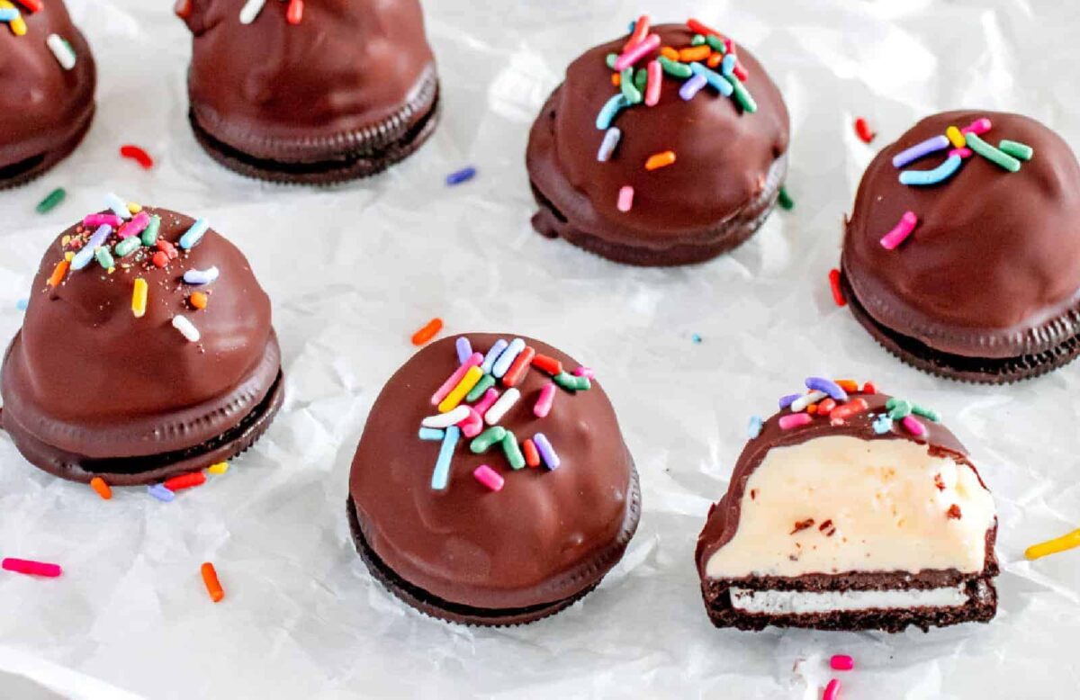 Chocolate-covered sandwich cookies topped with colorful sprinkles, with one cookie cut in half revealing a creamy filling inside—perfect for fans of ice cream desserts and other sweet treats.