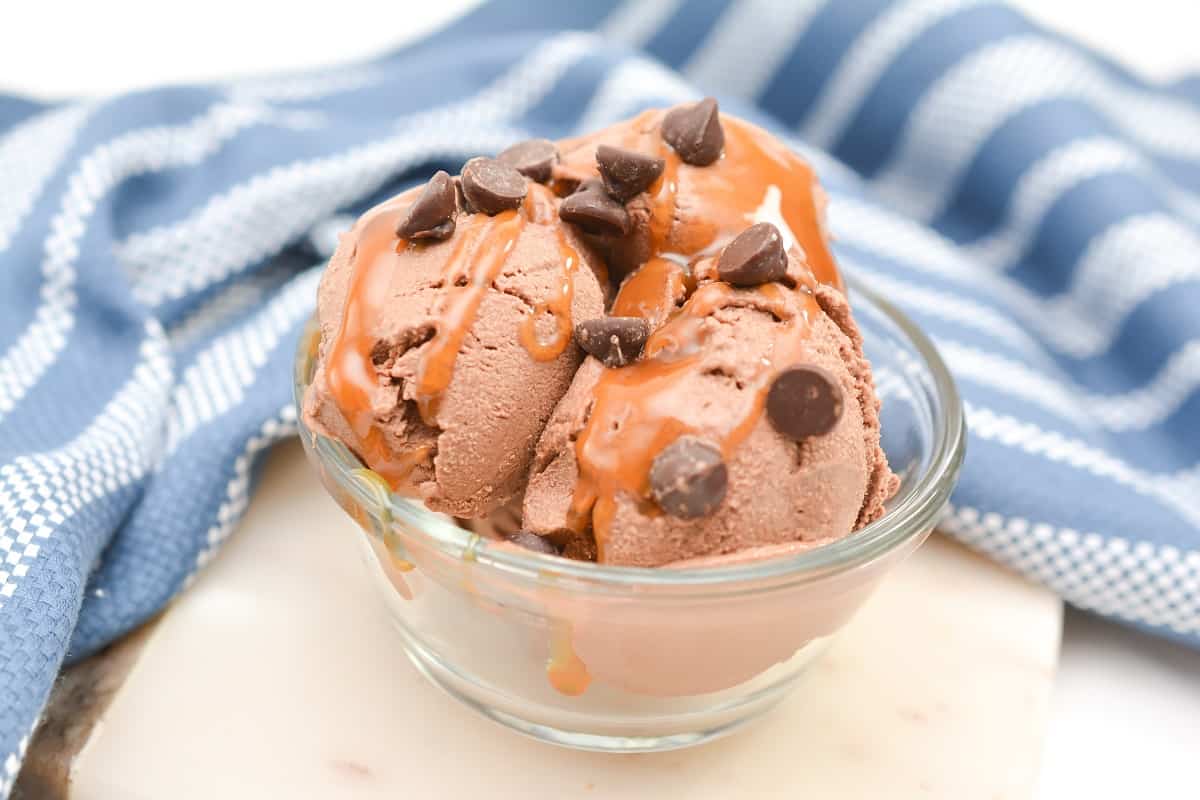 A glass bowl of chocolate ice cream, one of the classic ice cream desserts, topped with chocolate chips and caramel sauce, sits in front of a blue and white striped cloth.