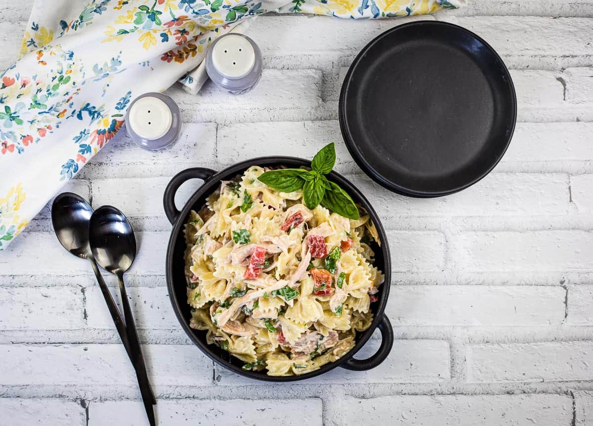 A bowl of kid-friendly pasta salad with vegetables and herbs sits on a white brick surface next to a black plate, two spoons, and salt and pepper shakers—perfect for easy pasta dinners.