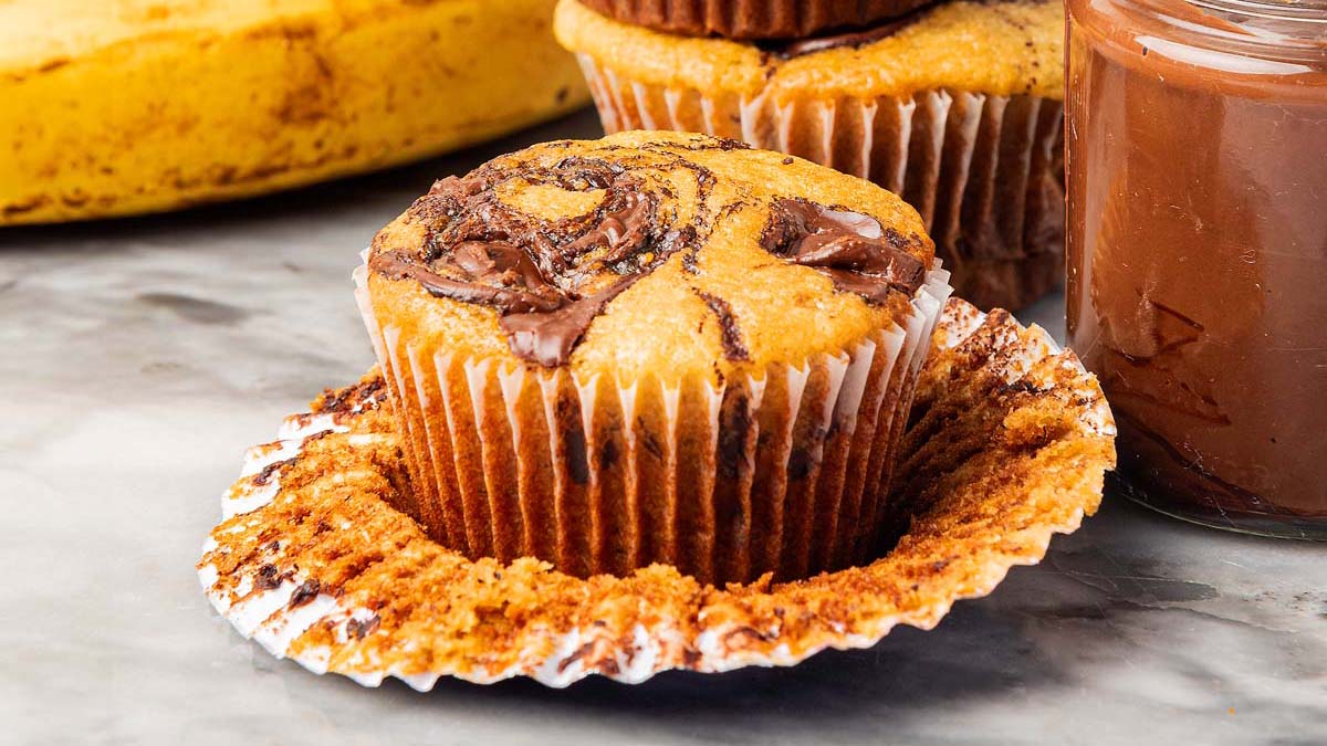 A close-up of a banana muffin with a Nutella swirl, partially unwrapped from its paper liner, sits on a marble surface next to a jar of chocolate spread—perfect inspiration for Nutella desserts.