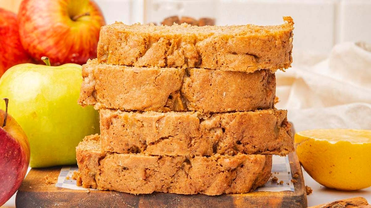 Four slices of apple bread stacked on a wooden board, surrounded by whole apples and a half lemon in the background—a perfect treat for loaf cake lovers craving big flavor.