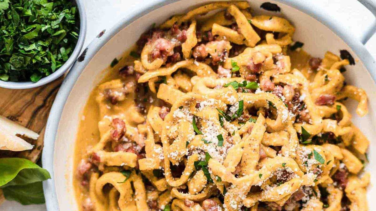 A plate of pasta topped with grated cheese, creamy sauce, bacon pieces, and chopped parsley—perfect for cozy evenings and inspiring new pasta recipes—with a bowl of fresh herbs on the side.