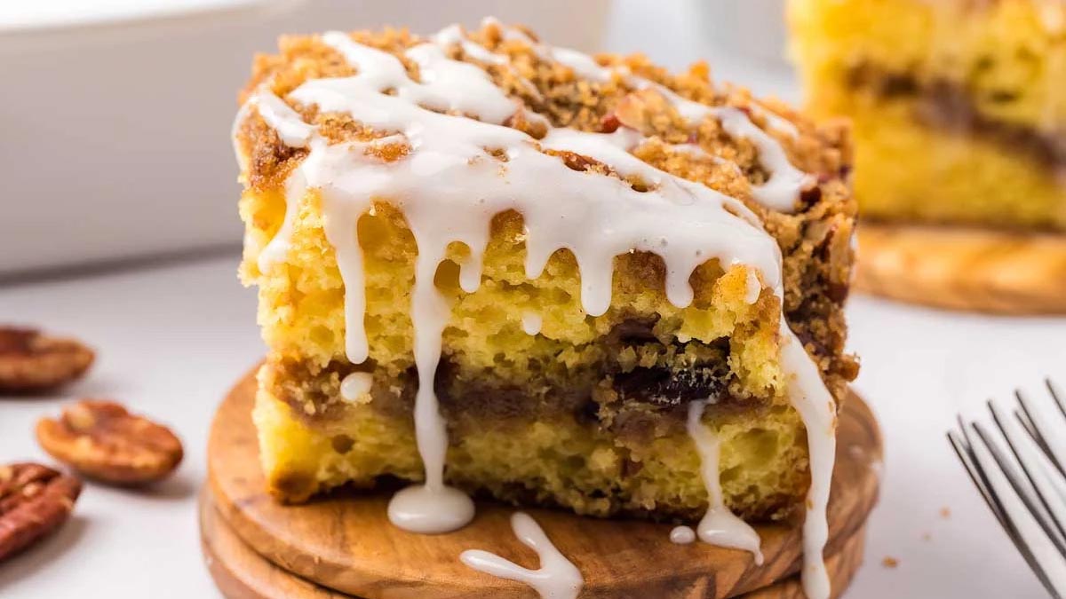 A slice of coffee cake with crumbly topping and white icing sits on a wooden coaster, with pecans and a fork nearby—perfect for those who love Coffee Cakes with soft centers.