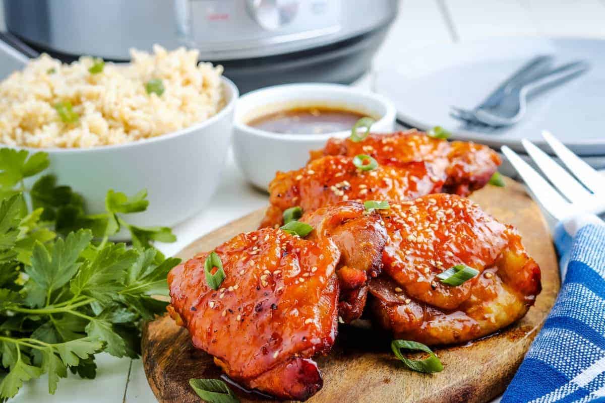 Three glazed chicken thighs garnished with chopped green onions rest on a wooden board, making for perfect quick dinners. Served with rice, sauce, fresh parsley, and utensils in the background for an easy recipe everyone will love.
