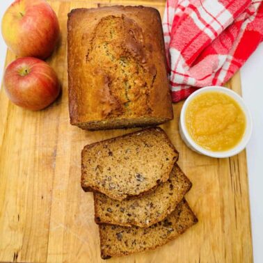 A loaf of sliced banana bread on a wooden board with two apples, a small bowl of applesauce, and a red checked kitchen towel—perfect for breakfast or as one of your favorite quick breads snacks.