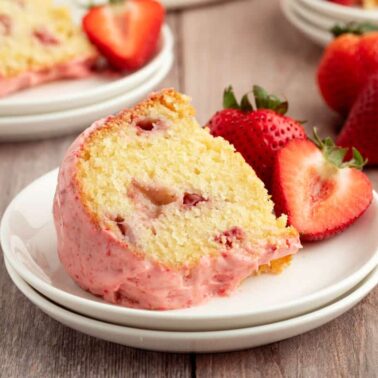 A slice of strawberry cake with pink icing on a white plate, garnished with fresh strawberries, brings classic flavors to life on a wooden table.