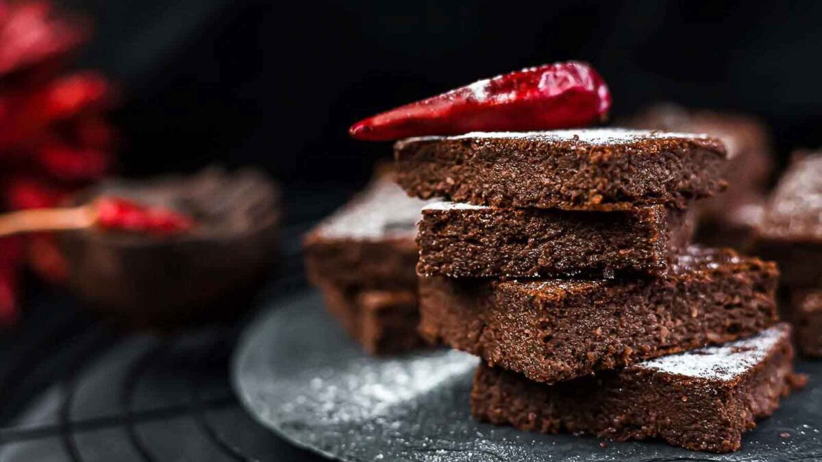 A stack of chocolate brownies dusted with powdered sugar, topped with a red chili pepper, sits on a dark plate—perfect desserts for those who crave unique after-school treats, with more brownies tempting in the background.
