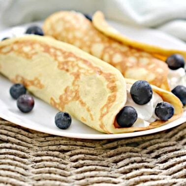 Two rolled crepes filled with cream and blueberries are served on a white plate, with extra blueberries scattered around—perfect for breakfast ideas and delicious morning meals.