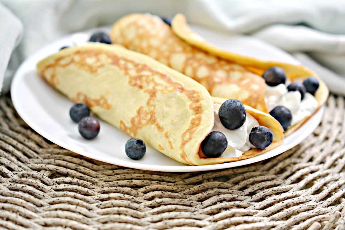 Two rolled crepes filled with cream and blueberries are served on a white plate, with extra blueberries scattered around—perfect for breakfast ideas and delicious morning meals.