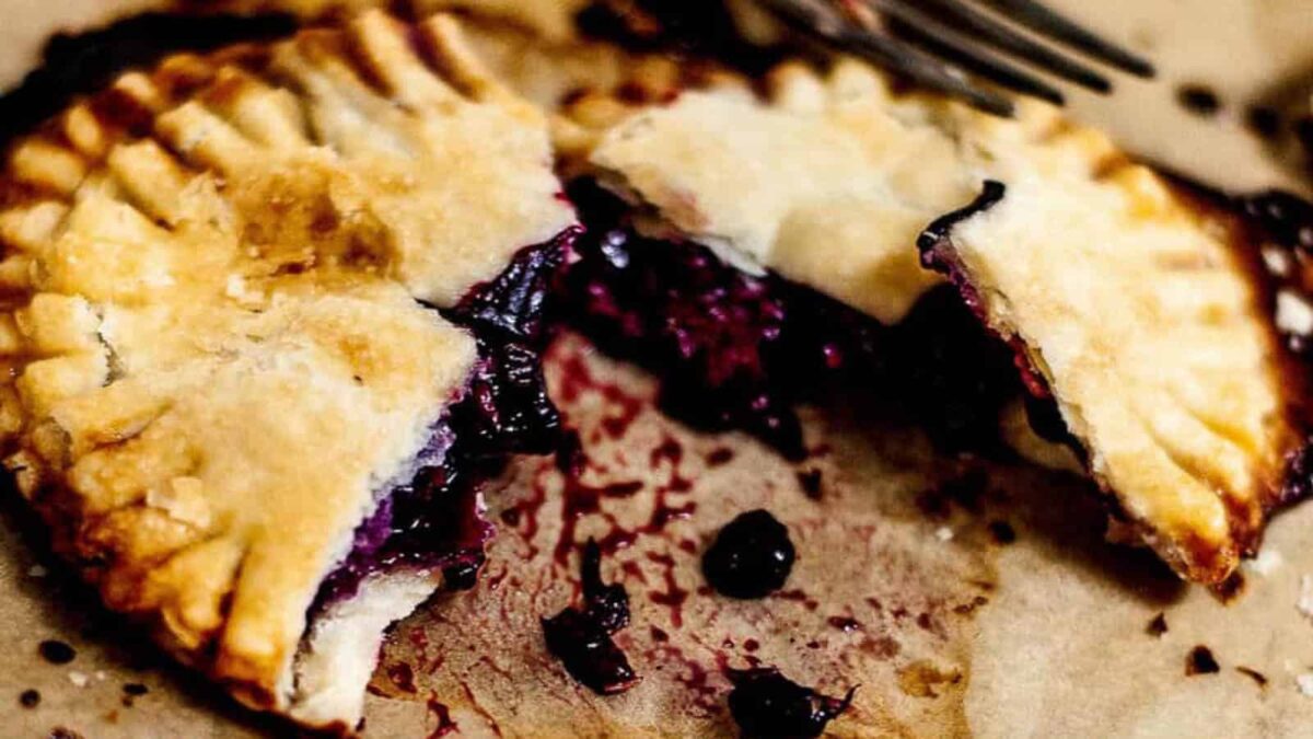 A baked hand pie with a flaky golden crust, broken in half to reveal a dark purple berry filling, sits on parchment paper with a fork nearby—one of the best kid-friendly treats for easy dessert recipes or after-school desserts.