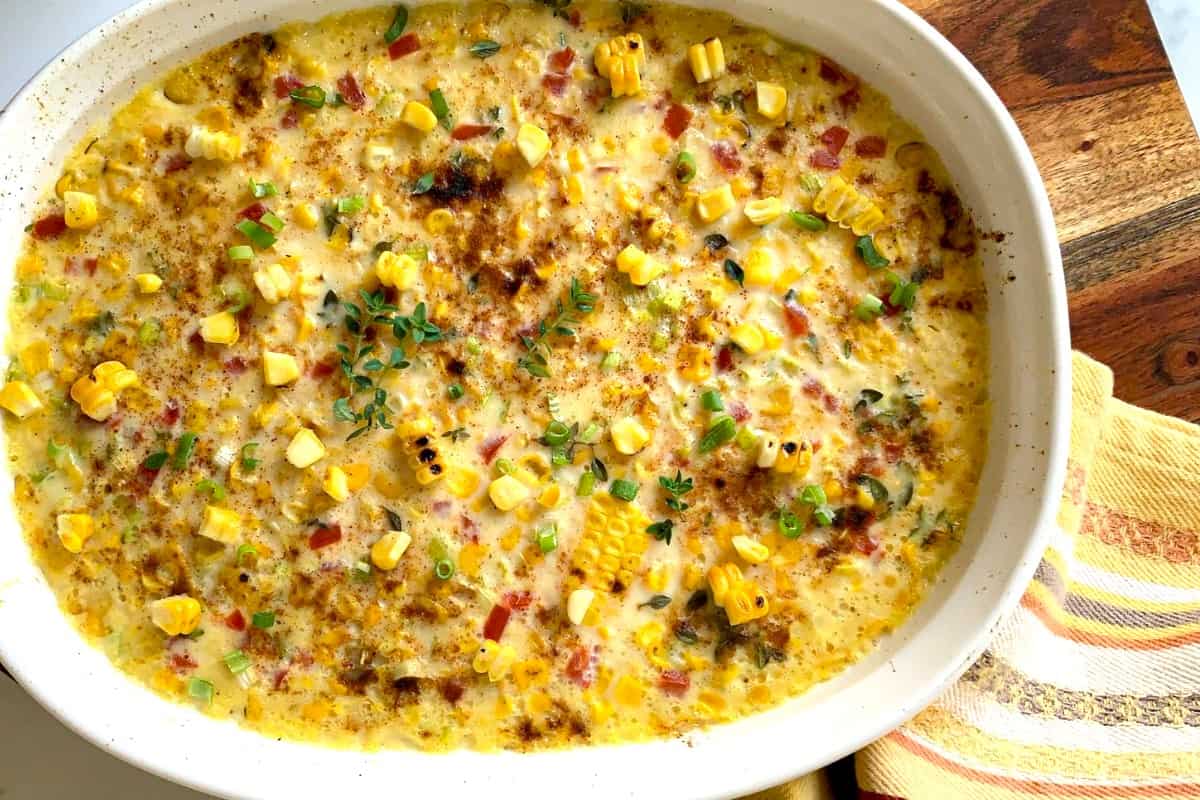A baked casserole dish filled with creamy corn pudding, topped with chopped green onions, herbs, and grilled corn pieces—one of the best fresh corn recipes for summer. Viewed from above.