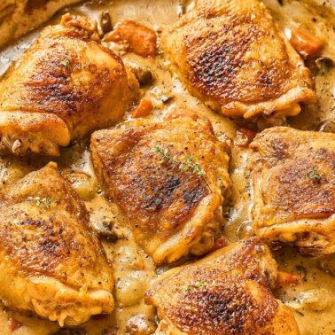 Six browned chicken thighs simmer in a creamy sauce with herbs and vegetables in a large skillet—perfect for delicious chicken dinners or anyone seeking easy chicken recipes.