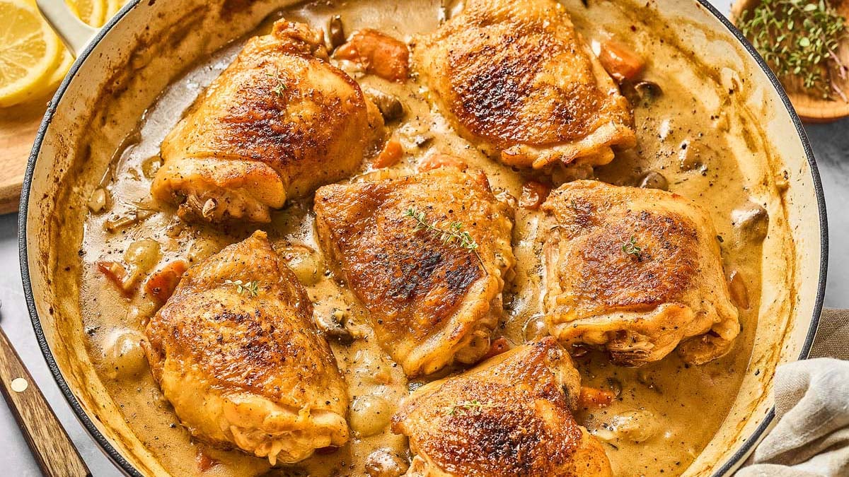 Six browned chicken thighs simmer in a creamy sauce with herbs and vegetables in a large skillet—perfect for delicious chicken dinners or anyone seeking easy chicken recipes.