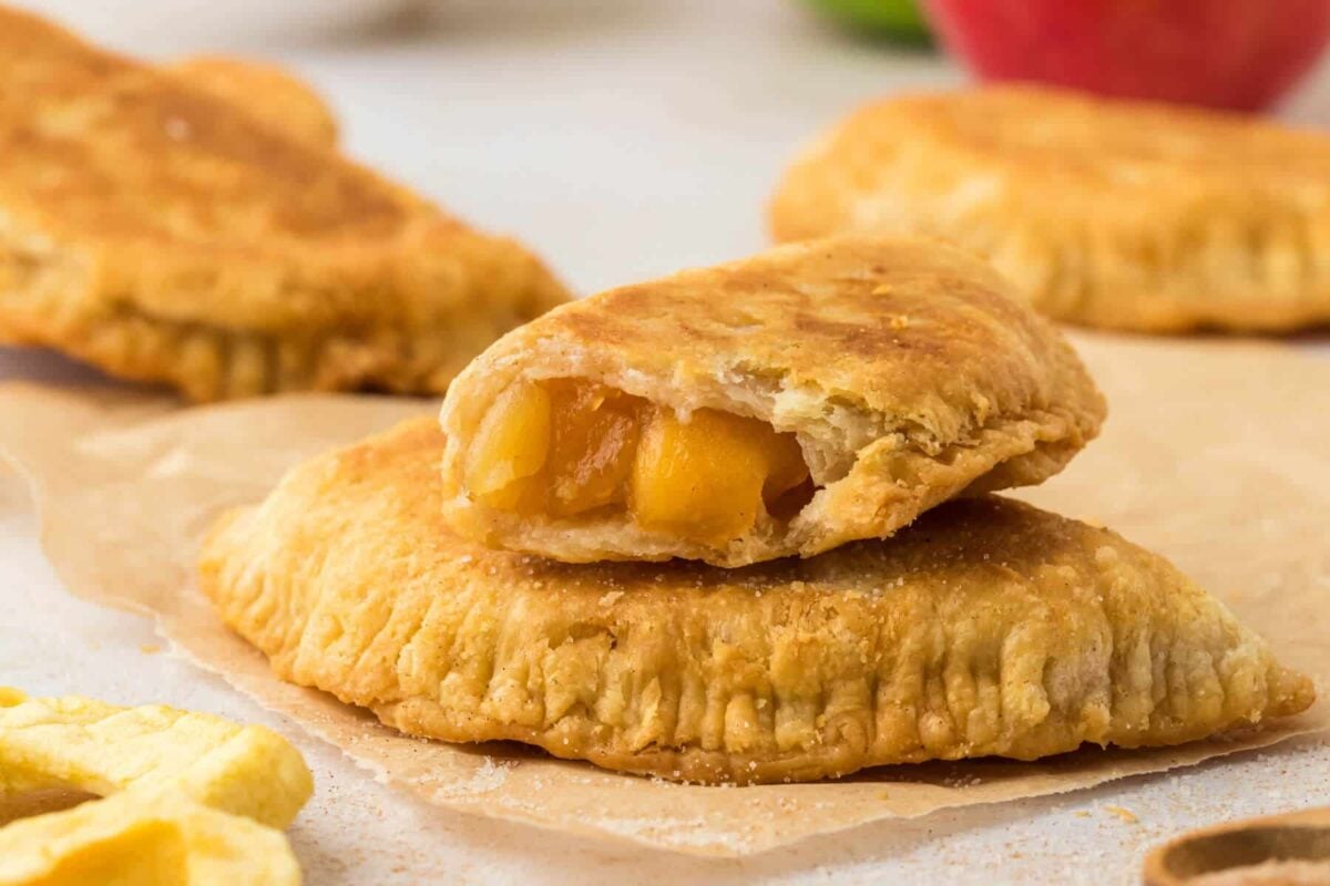 A golden-brown fried hand pie is shown with one half broken open, revealing a warm apple filling. These creative apple pie treats join other hand pies in the background on parchment paper, perfect for fans of unique Apple Pie Recipes.