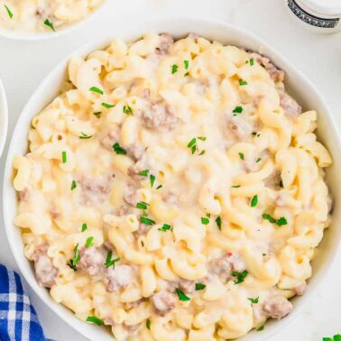 A bowl of creamy macaroni and cheese with ground meat, garnished with chopped parsley, sits on a white surface next to smaller bowls, salt and pepper shakers, fresh parsley, and hints of decadent desserts for a truly comforting meal.