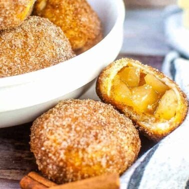 A bowl of sugar-coated dough balls sits beside a halved dough ball filled with apple pie filling—delicious vegan snacks. Cinnamon sticks and butter are also visible on the table, making these treats perfect for those seeking tasty vegan recipes.