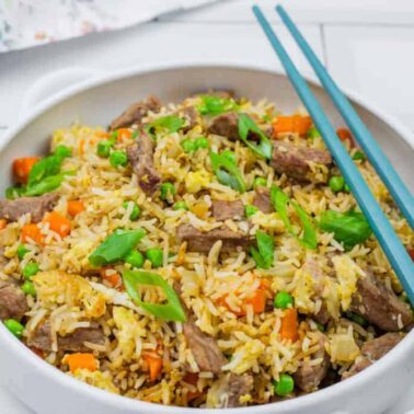 A bowl of beef fried rice with peas, carrots, green onions, and blue chopsticks placed on the rim—one of those epic rice recipes perfect for fans of easy, cheap meals.