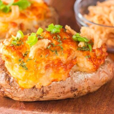 Baked potatoes topped with melted cheese, green onions, and crispy fried onions make one of the best air fryer side dishes, served on a wooden surface next to a bowl of fried onions and a red plaid napkin.
