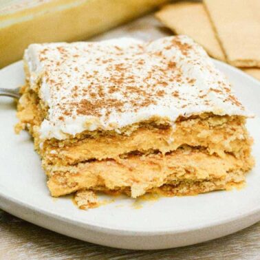 A square slice of layered pumpkin dessert with a graham cracker base and whipped topping, sprinkled with cinnamon, served on a white plate—a delightful choice for autumn desserts.