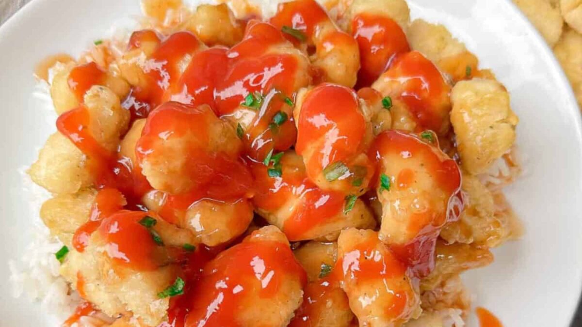 A plate of white rice topped with battered, fried chicken pieces covered in a red sweet and sour sauce, garnished with chopped green herbs—a cozy choice for fall dinners and perfect for sweater weather.