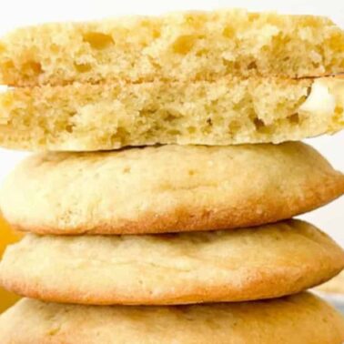 A stack of three low carb cookies with a fourth cookie on top, broken in half to reveal the soft, moist interior. Lemons are visible in the background.