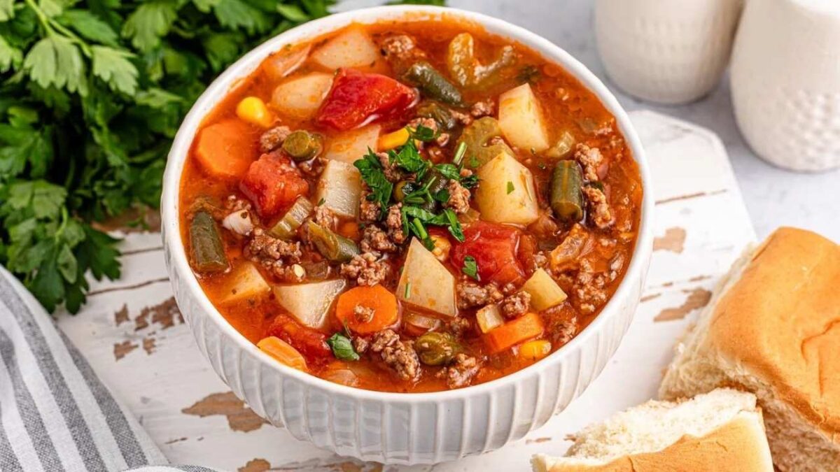 A bowl of hearty vegetable and beef soup with potatoes, carrots, green beans, and tomatoes, garnished with herbs—one of our favorite easy soups—served with a dinner roll on the side.
