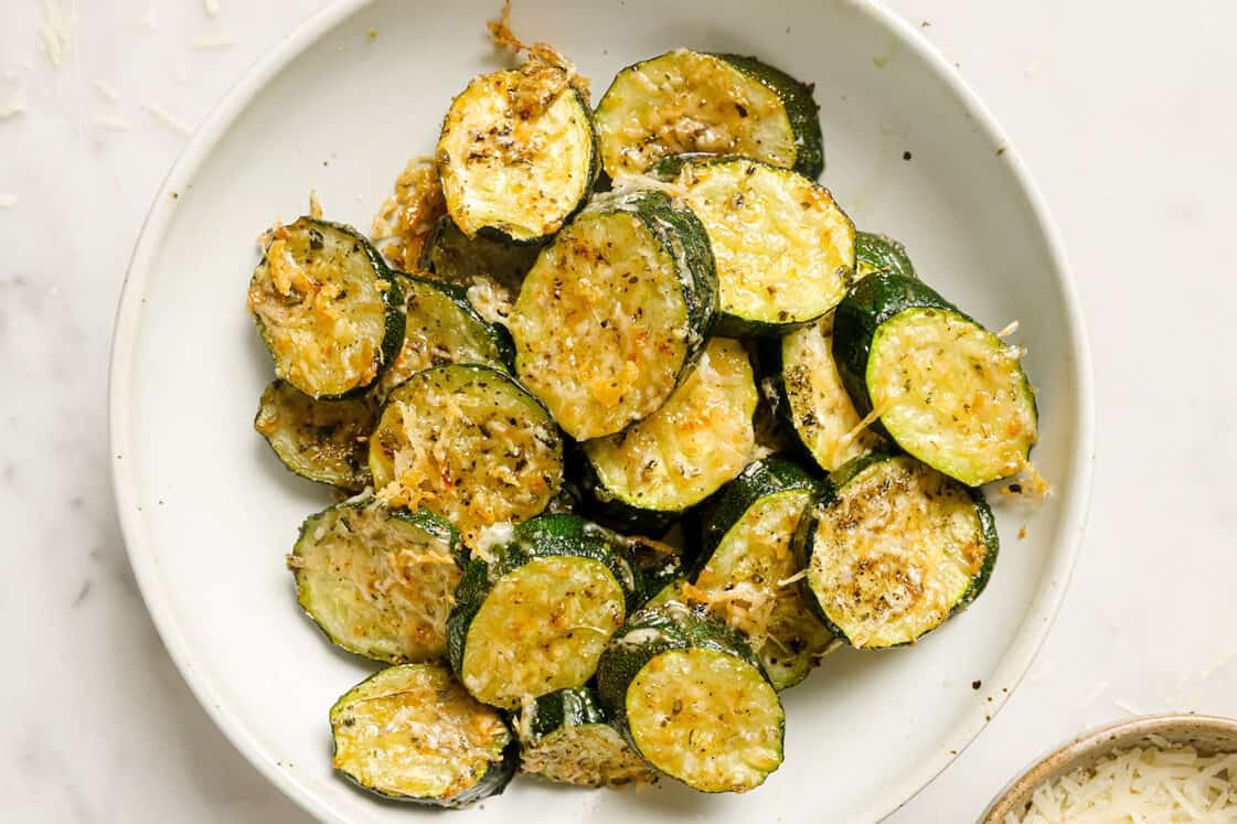 A white plate filled with roasted zucchini rounds topped with melted cheese and herbs—one of those delicious side dishes that easily steals the show at any main event.