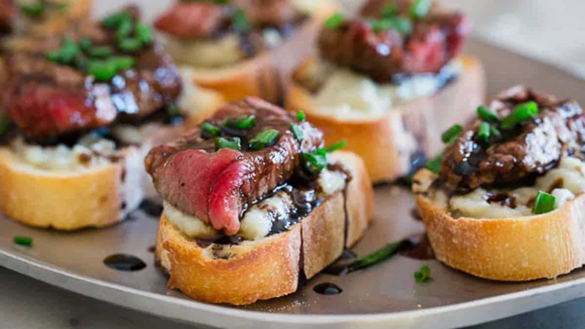Slices of toasted baguette topped with mashed potatoes, steak pieces, balsamic glaze, and chopped chives, arranged on a tray—a perfect choice for quick meals or easy dinner recipes.