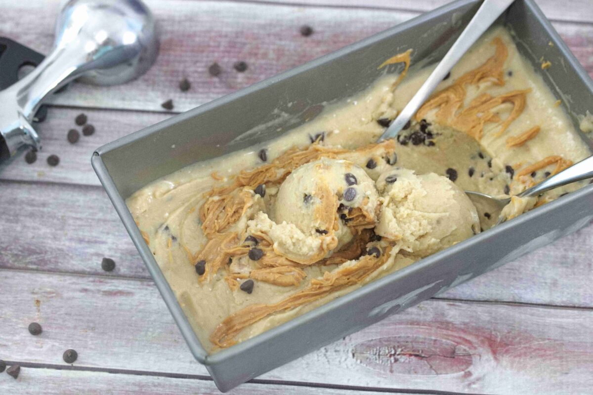 A rectangular metal container holds peanut butter chocolate chip ice cream with swirls of peanut butter, two scoops, and a spoon. An ice cream scooper rests nearby on a wooden surface—perfect for fans of easy, oven-free desserts.