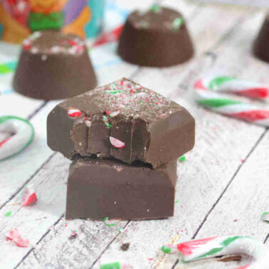 Two chocolate candies, one with a bite taken out, are stacked among broken pieces of candy canes on a white wooden surface—perfect inspiration for easy desserts and festive no-bake recipes.