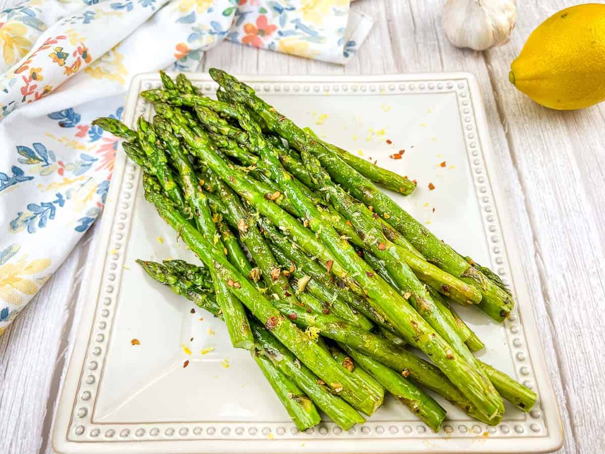 A square white plate with cooked asparagus spears, garnished with seasonings and lemon zest—perfect for summer recipes. A lemon, garlic, and a floral napkin are nearby on a light wooden surface.