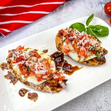 Two pieces of grilled chicken topped with mozzarella, diced tomato, basil, and onions, drizzled with balsamic glaze—perfect for summer recipes—served on a white plate with a sprig of basil and cherry tomatoes.