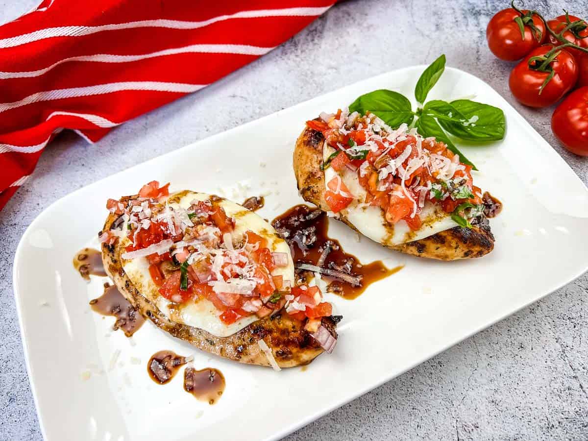Two pieces of grilled chicken topped with mozzarella, diced tomato, basil, and onions, drizzled with balsamic glaze—perfect for summer recipes—served on a white plate with a sprig of basil and cherry tomatoes.