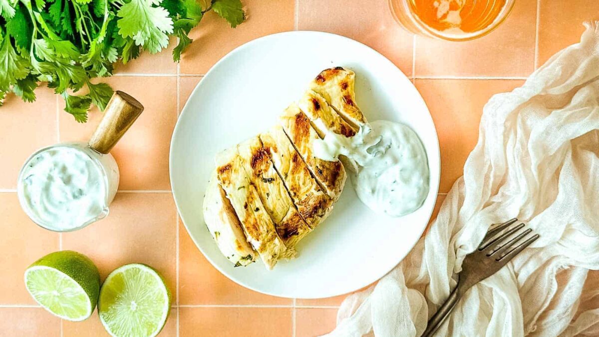 Grilled chicken breast with white sauce on a white plate, garnished with cilantro, lime halves, a glass of sauce, and a fork on a peach-colored tiled surface—a delicious addition to your summer recipes.