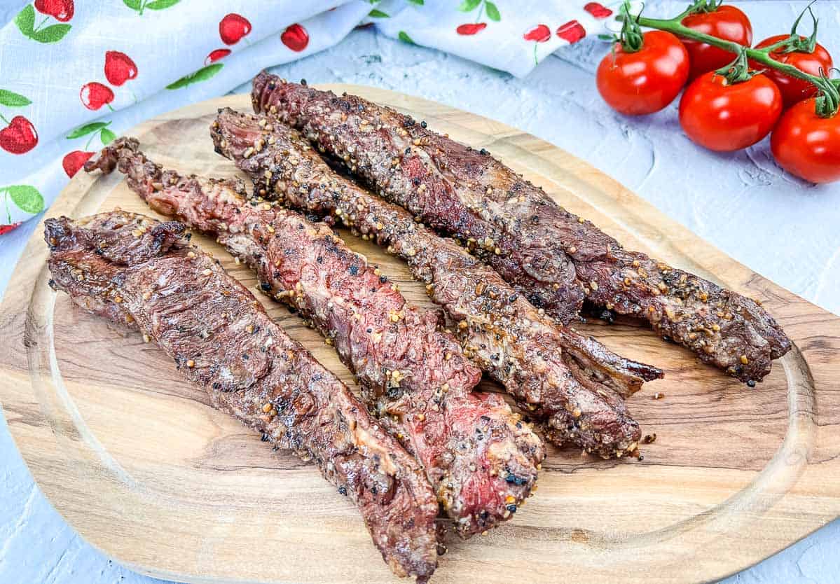 Four cooked, seasoned steak strips are arranged on a wooden cutting board beside fresh tomatoes and a cherry-patterned cloth—perfect inspiration for your next grilling ideas or summer recipes.