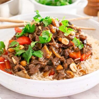 A bowl of rice topped with stir-fried beef, red bell pepper, onions, peanuts, and fresh cilantro—an ideal choice for simple meals with easy cleanup. Chopsticks rest on the bowl, ready for a quick and delicious experience.