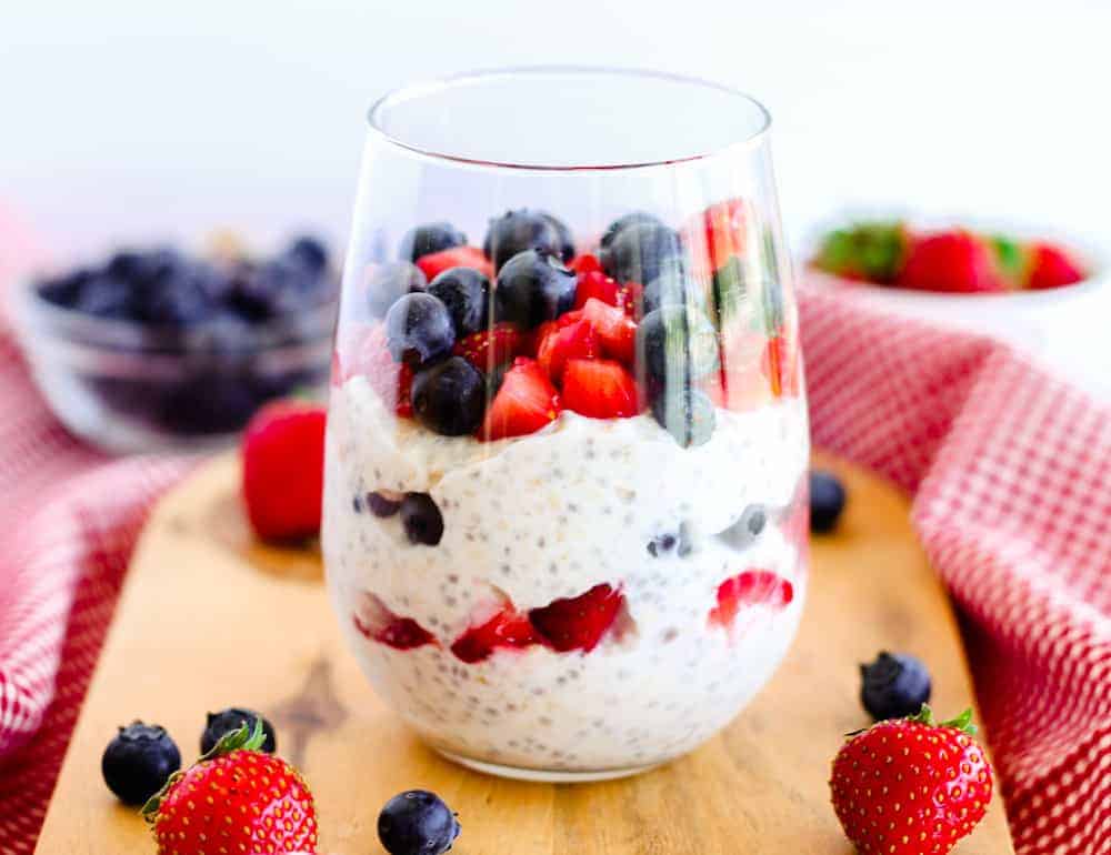 A glass filled with chia seed pudding layered with strawberries and blueberries, placed on a wooden board, offers exciting breakfast inspiration, surrounded by whole berries and a red cloth in the background.