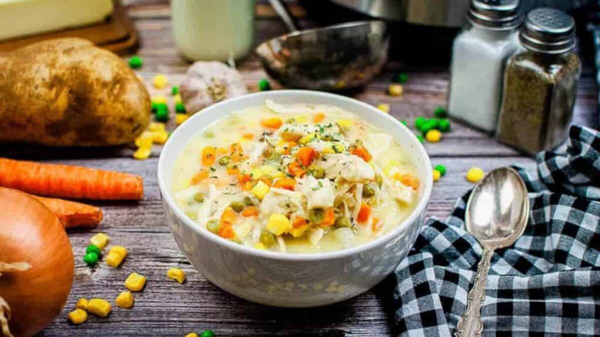 A bowl of creamy chicken vegetable soup, one of the best healing soups, is garnished with herbs and surrounded by carrots, onion, potato, a spoon, and scattered mixed vegetables on a wooden table.