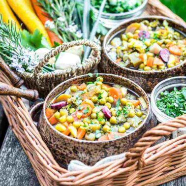 A wicker tray holding bowls of hearty vegetable stew inspired by healthy soups, with chopped herbs and colorful sliced carrots, set on a wooden outdoor table.